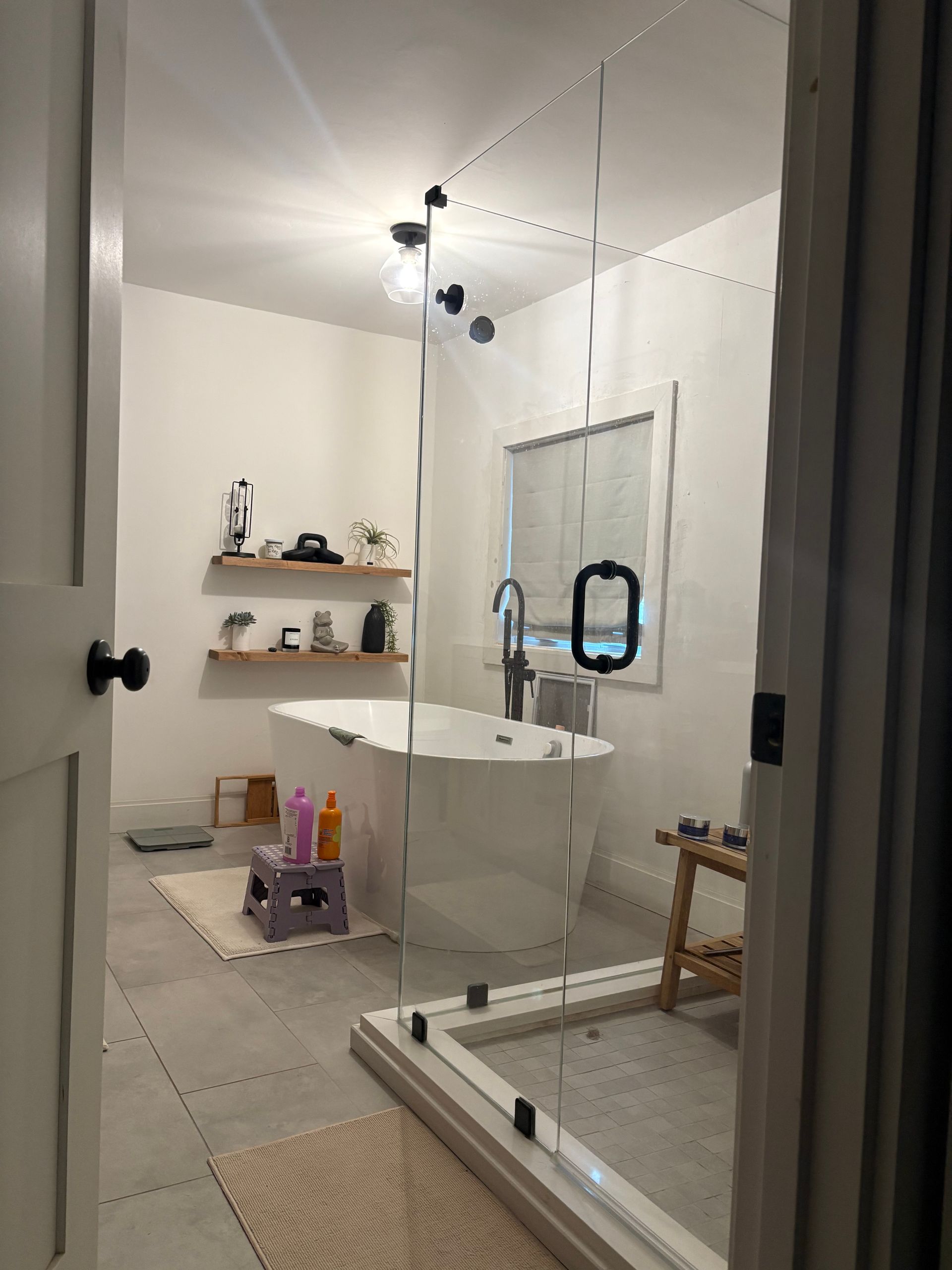 A bathroom viewed through a doorway, featuring a freestanding white bathtub, a glass-enclosed shower, and wood shelves.