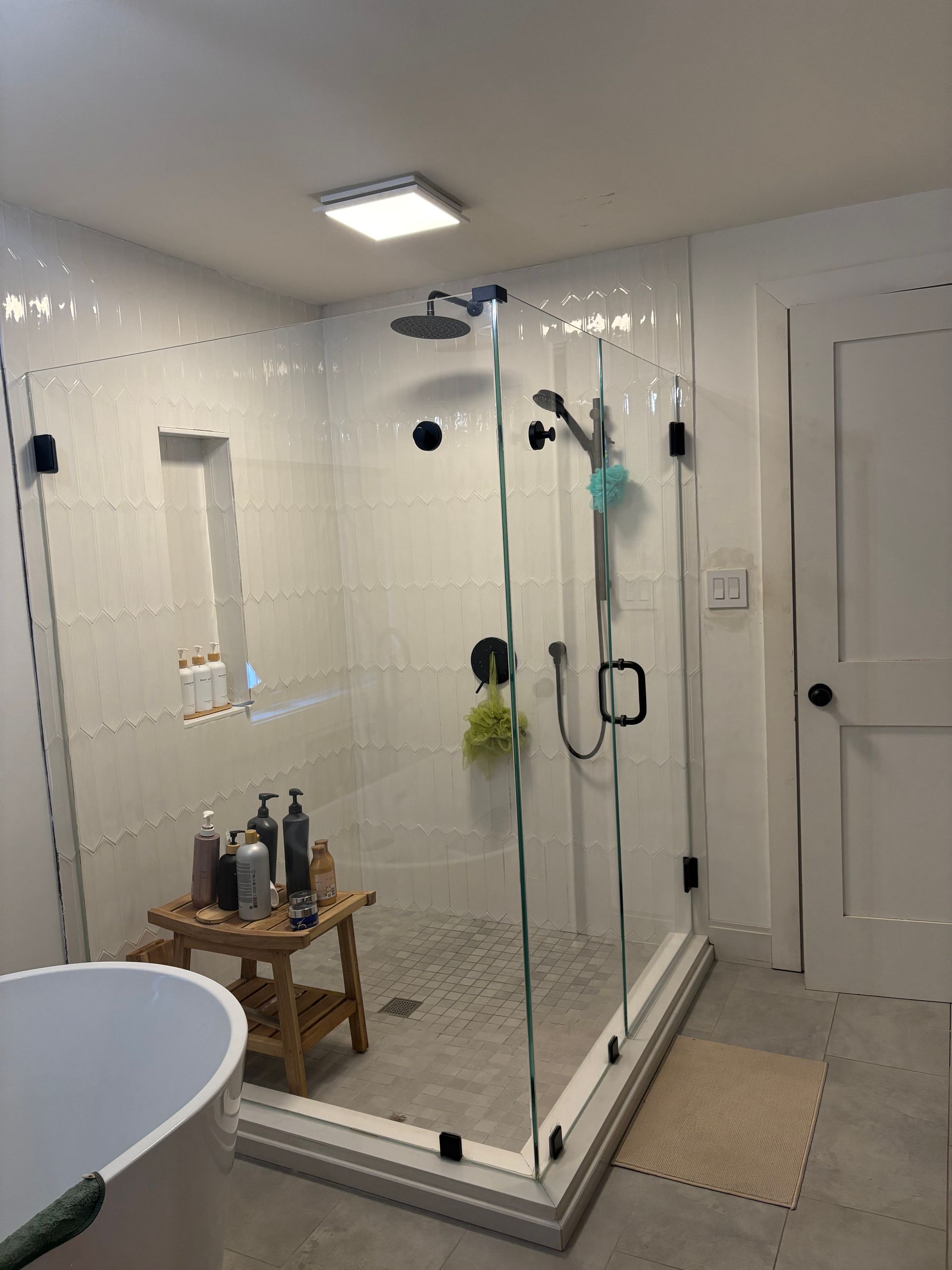 A modern walk-in shower with white tile, glass enclosure, black hardware, and a small wooden stool inside.