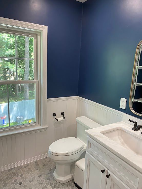 A bathroom with navy blue walls over white wainscoting, featuring a window, toilet, and vanity with a vanity mirror.