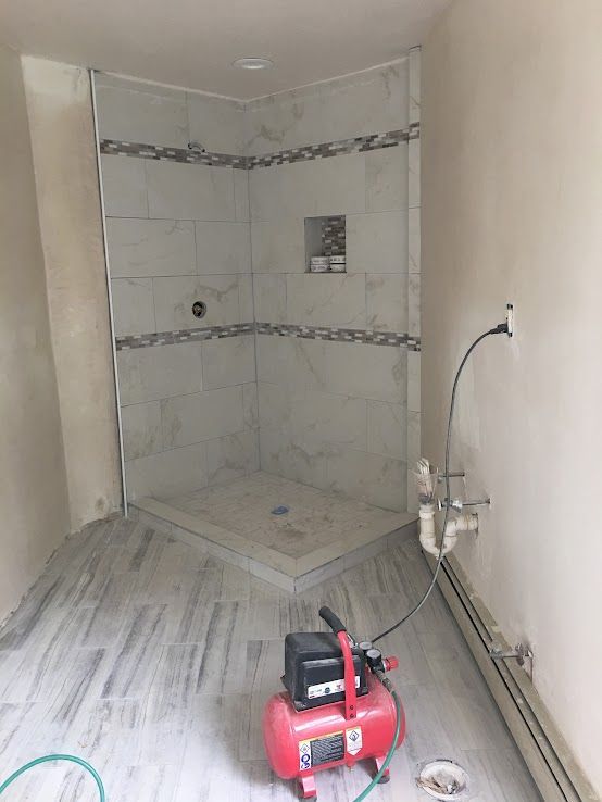 A bathroom under construction with a tiled corner shower, wood-look floor tiles, exposed plumbing, and an air compressor.