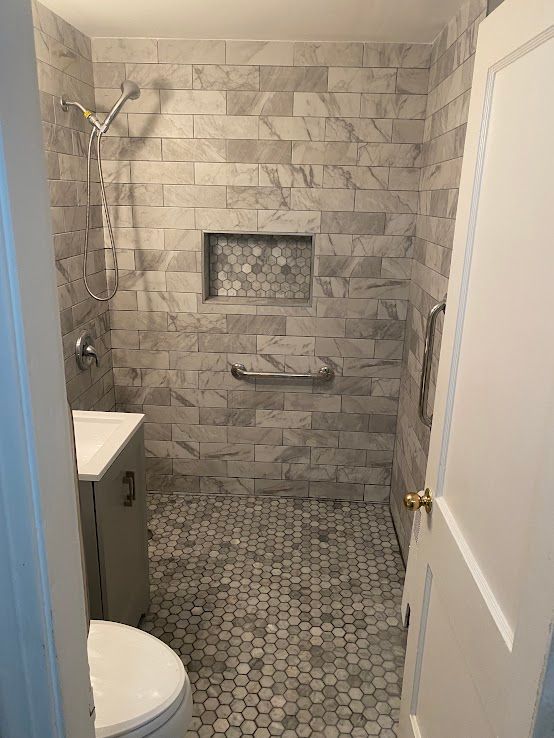 A walk-in shower with light grey stone walls, a hexagonal tiled floor, a built-in niche, and silver grab bars.