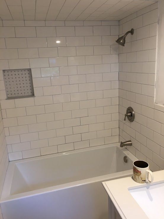 A white bathtub and shower surround with white subway tile and a small built-in shelf, with a mug sitting on a vanity.