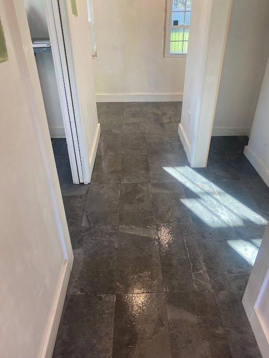 A high-angle view of a hallway featuring dark grey, textured stone tile flooring with bright sunlight casting shadows.