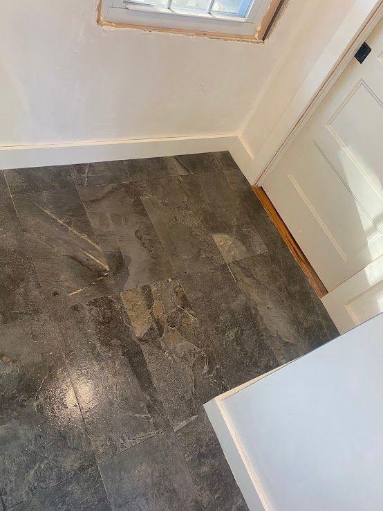 Dark grey, stone-look tile flooring in a room corner with white baseboards, a wall, and part of a door frame.