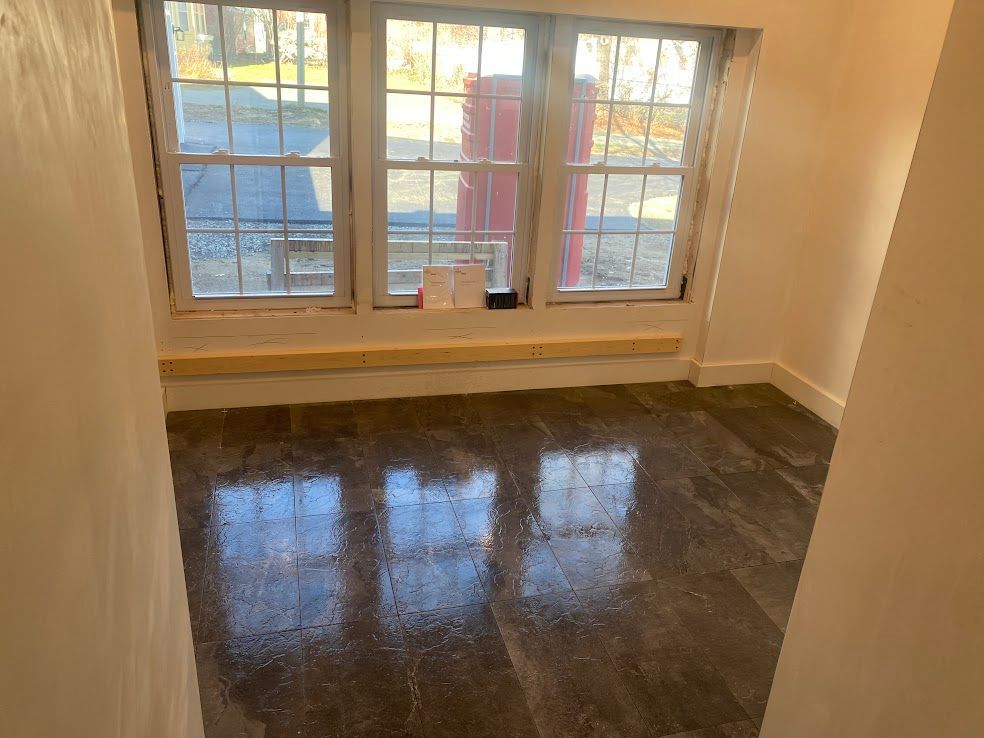 A view of a small, empty room with grey tiled flooring, white walls, and a large triple window with a wooden base.