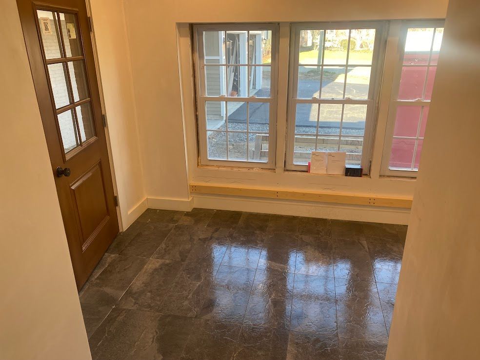 A small room with a wooden door, dark stone-look tile flooring, and three windows looking out toward a paved driveway.