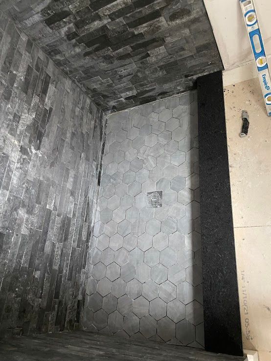 A shower under construction with dark stone tile walls, a gray hexagonal tile floor, and a black bench seat.