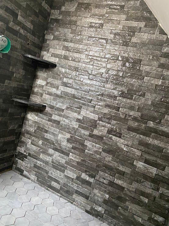 A shower interior with dark, textured stone-look wall tiles, two built-in corner shelves, and light-colored hexagonal floor.