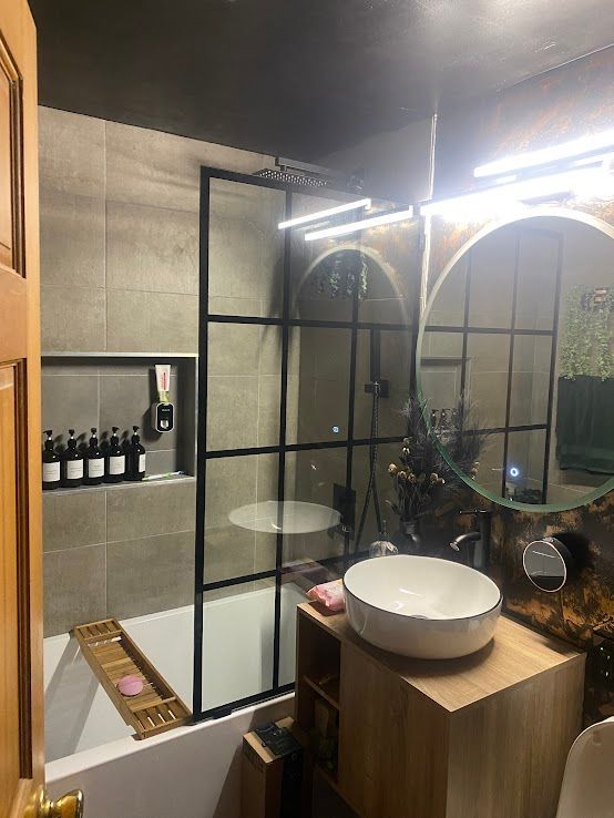 A modern bathroom featuring a white vessel sink, wooden vanity, and a black-framed glass shower screen over a bathtub.