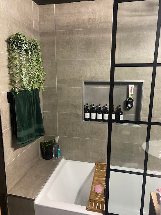 A modern bathroom featuring a white tub, a recessed shelf filled with bottles, a hanging plant, and a glass partition.