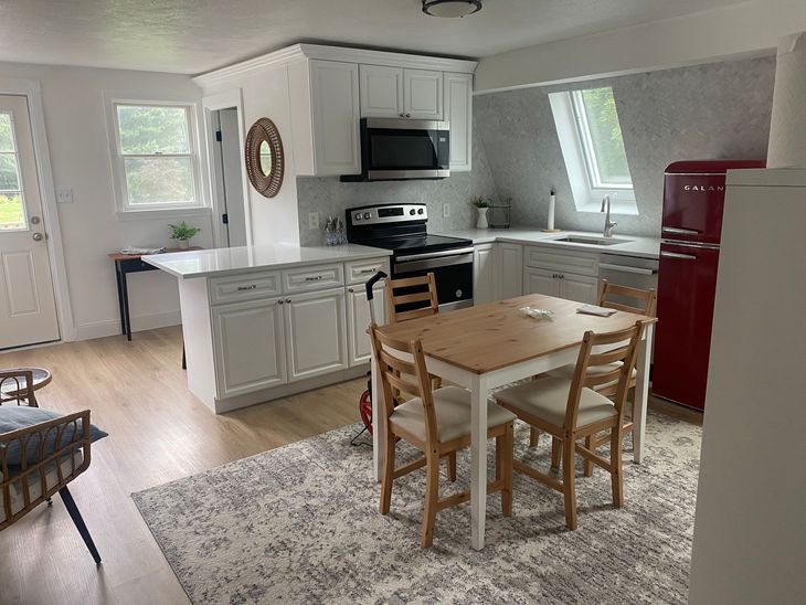 A bright kitchen with white cabinets, a red refrigerator, a wooden dining table with four chairs, and a patterned rug.