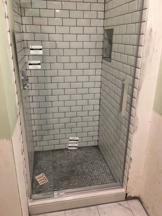 A walk-in shower with white subway tile walls, a gray mosaic tile floor, and a glass door, currently under construction.