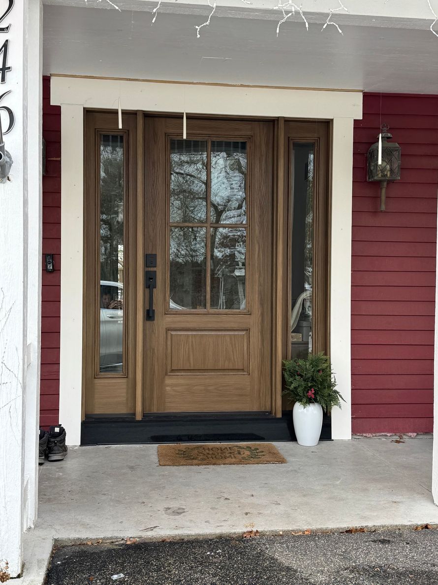 A wooden front door with glass panels and matching sidelights set against red siding, with a white frame and a potted plant.