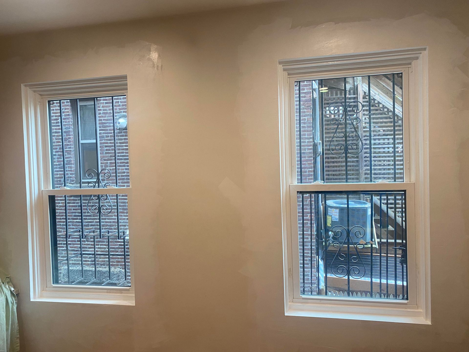 Two white-trimmed windows set into a beige wall, reflecting the brick building exterior and fire escape outside.