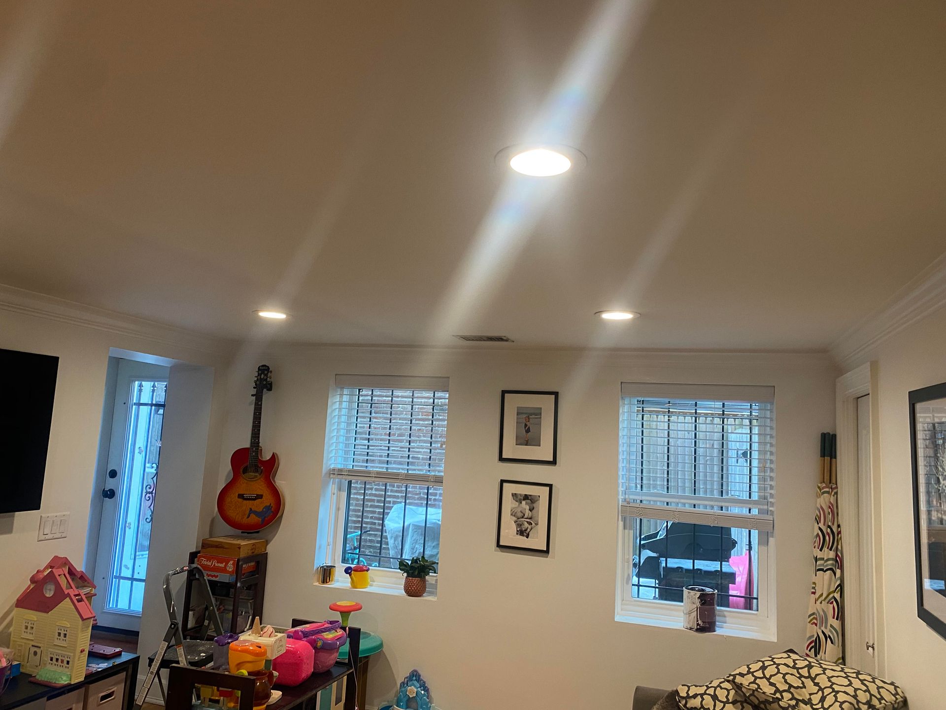 A room with two windows, a guitar on the wall, framed art, and various items on a table under recessed ceiling lights.
