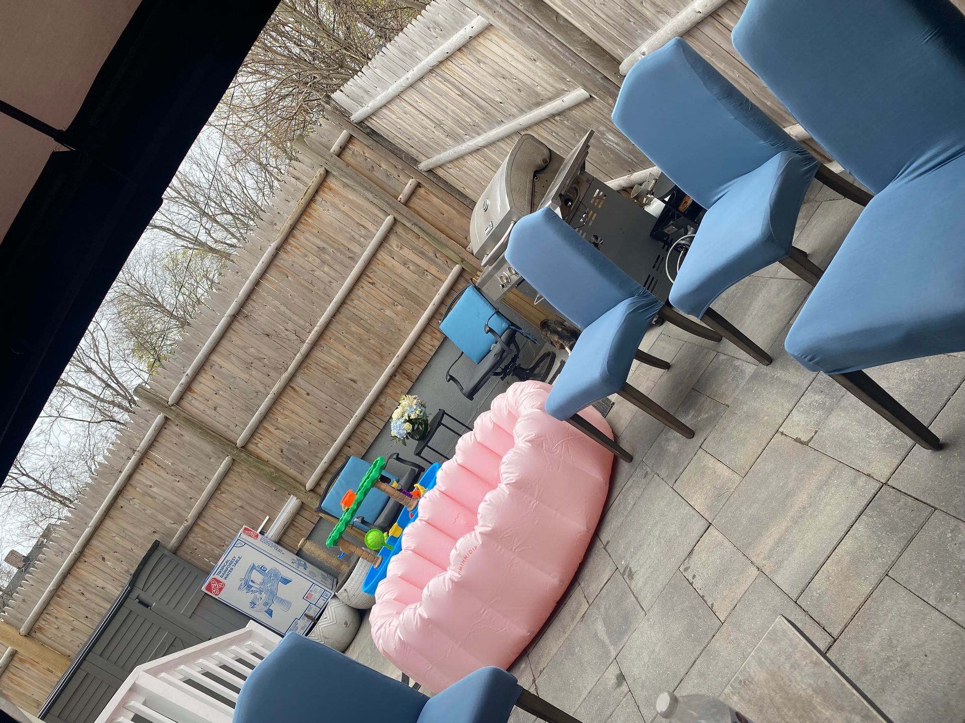 A pink inflatable pool sits on a stone patio near several blue chairs and a wooden fence.