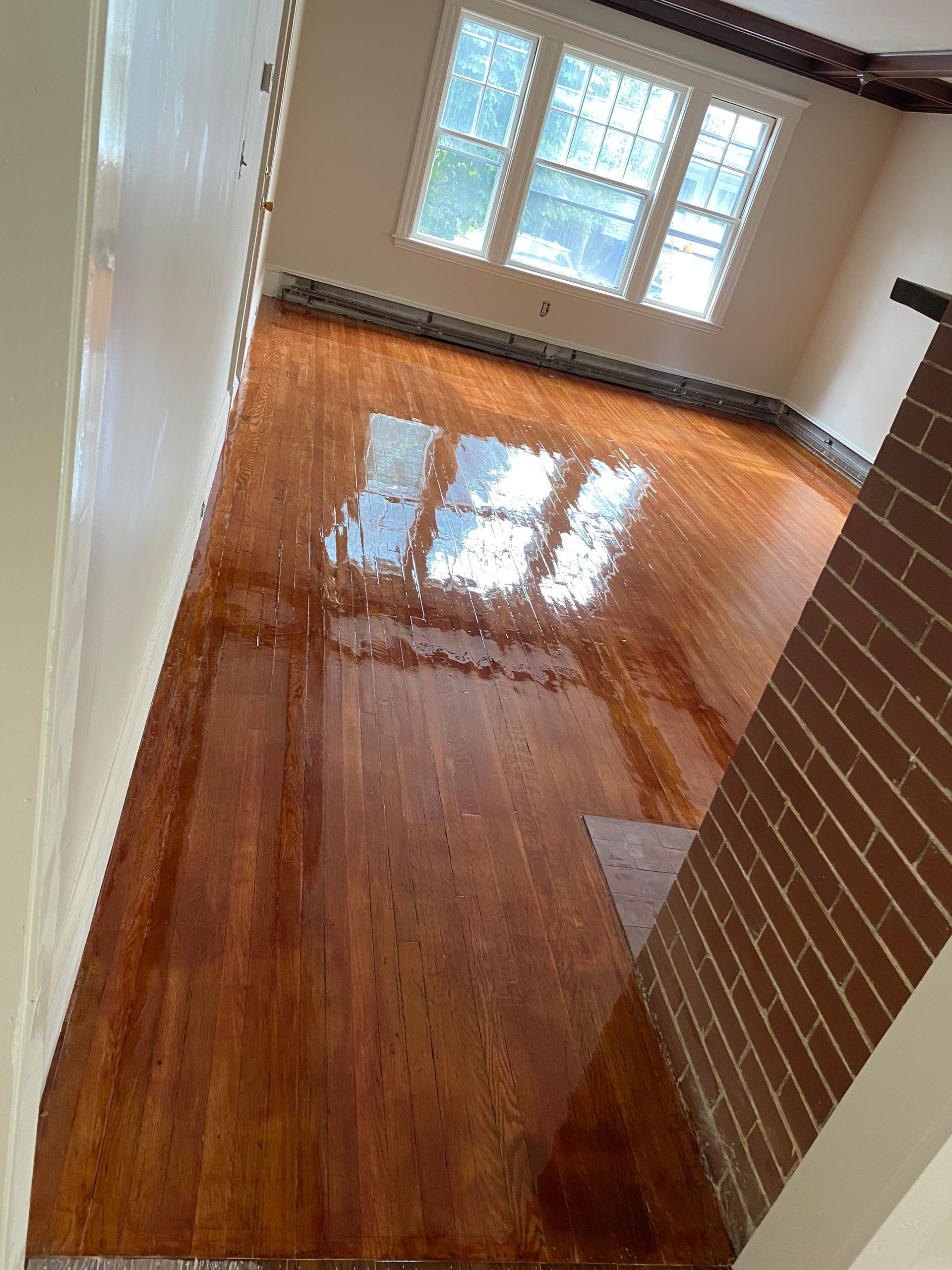 A room with polished, light-brown wooden floors reflecting the windows, adjacent to a brick wall and cream-colored walls.