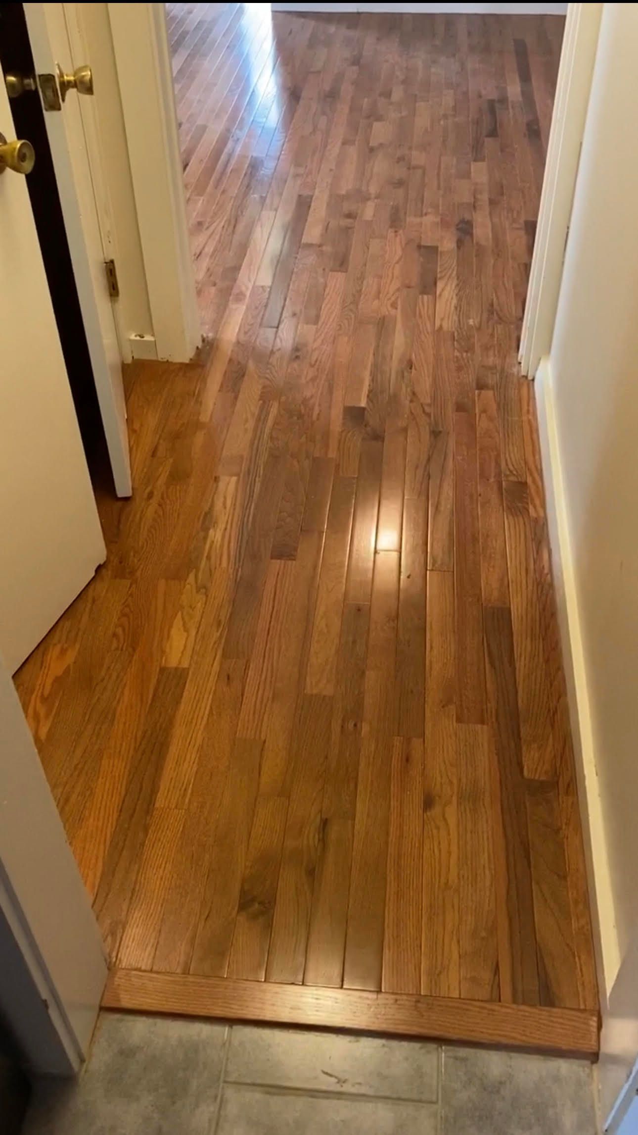 A hallway with light-brown hardwood flooring extending into an adjacent room, separated by a doorway.
