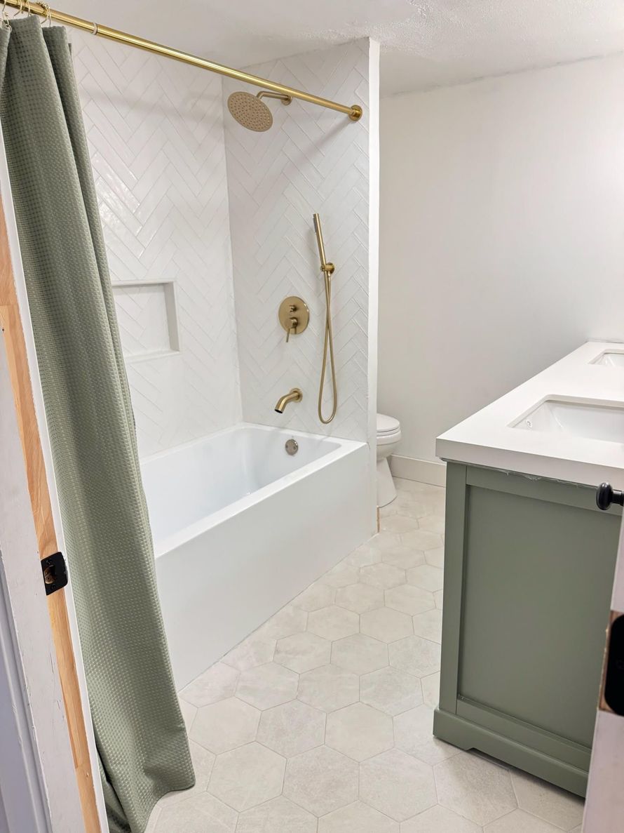 A modern bathroom with a white herringbone tiled tub, gold fixtures, a sage green vanity, and light patterned flooring.