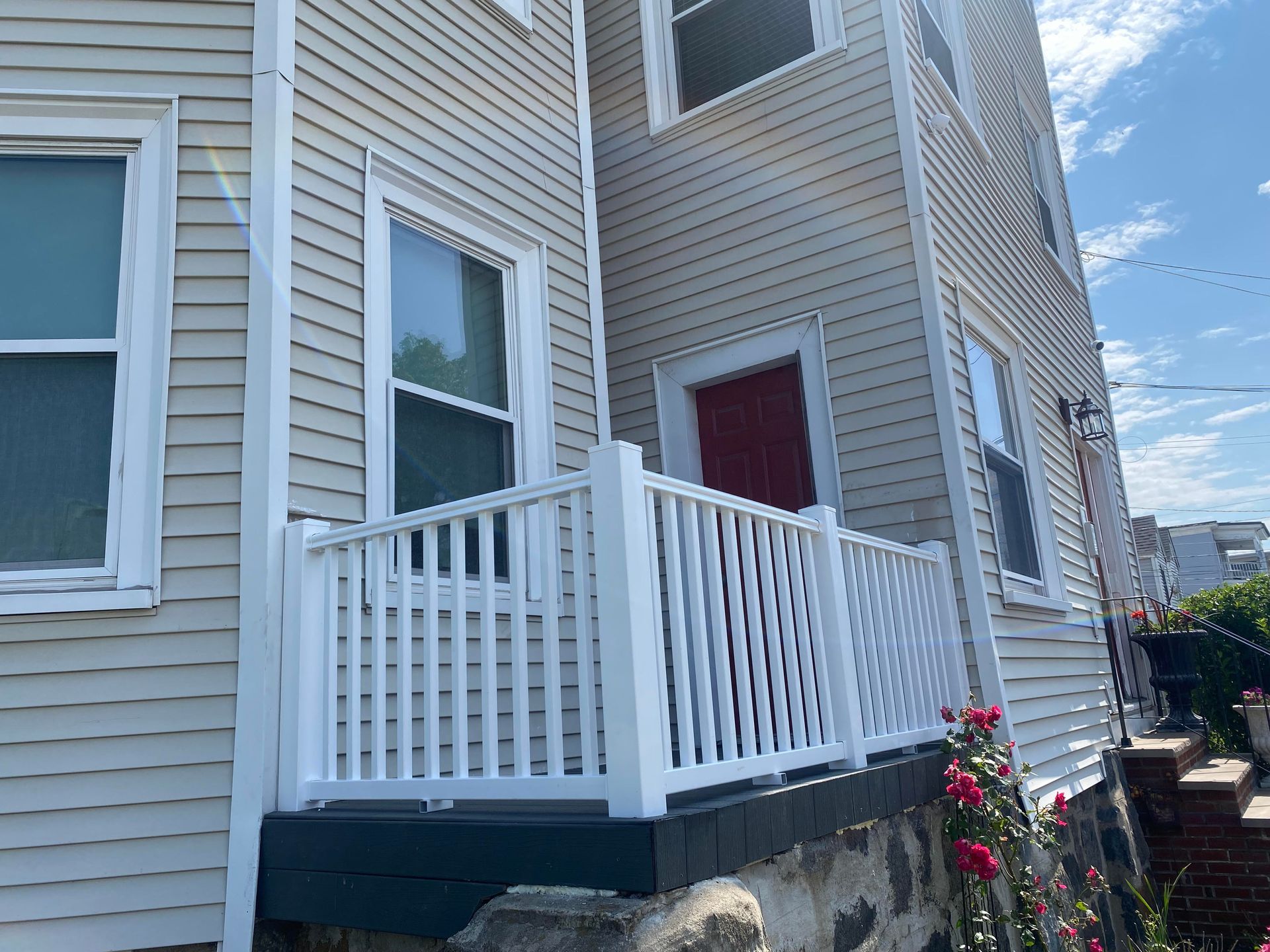A white wooden porch railing sits in front of a tan-sided house with a red door, stone foundation, and rose bushes.