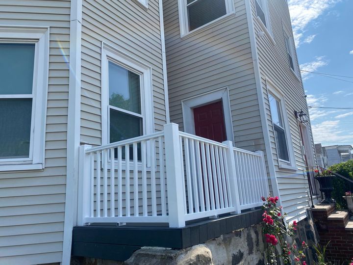 Light beige, multi-story house with a red door, white vinyl porch railing, and a stone foundation on a sunny day.