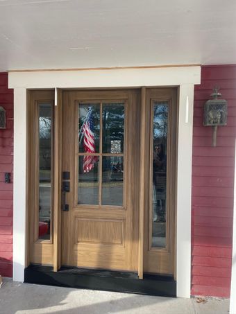A wooden front door with a glass window and side panels, framed in white trim, set against red exterior siding.