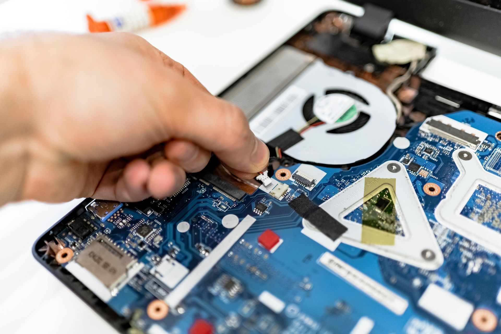 A hand carefully disconnects a small ribbon cable from the blue motherboard inside an open laptop.