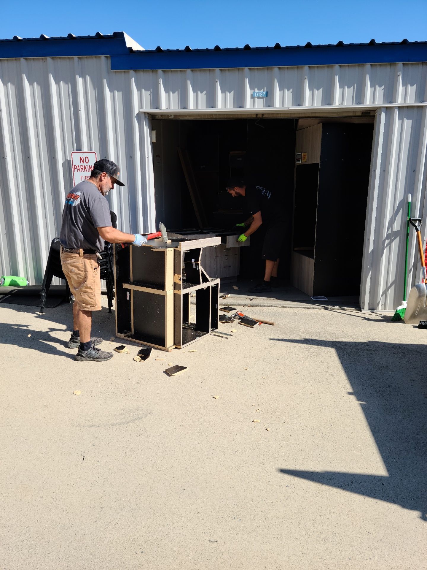 Two people working at a table saw in an open storage unit. One is cutting wood, the other is inside.