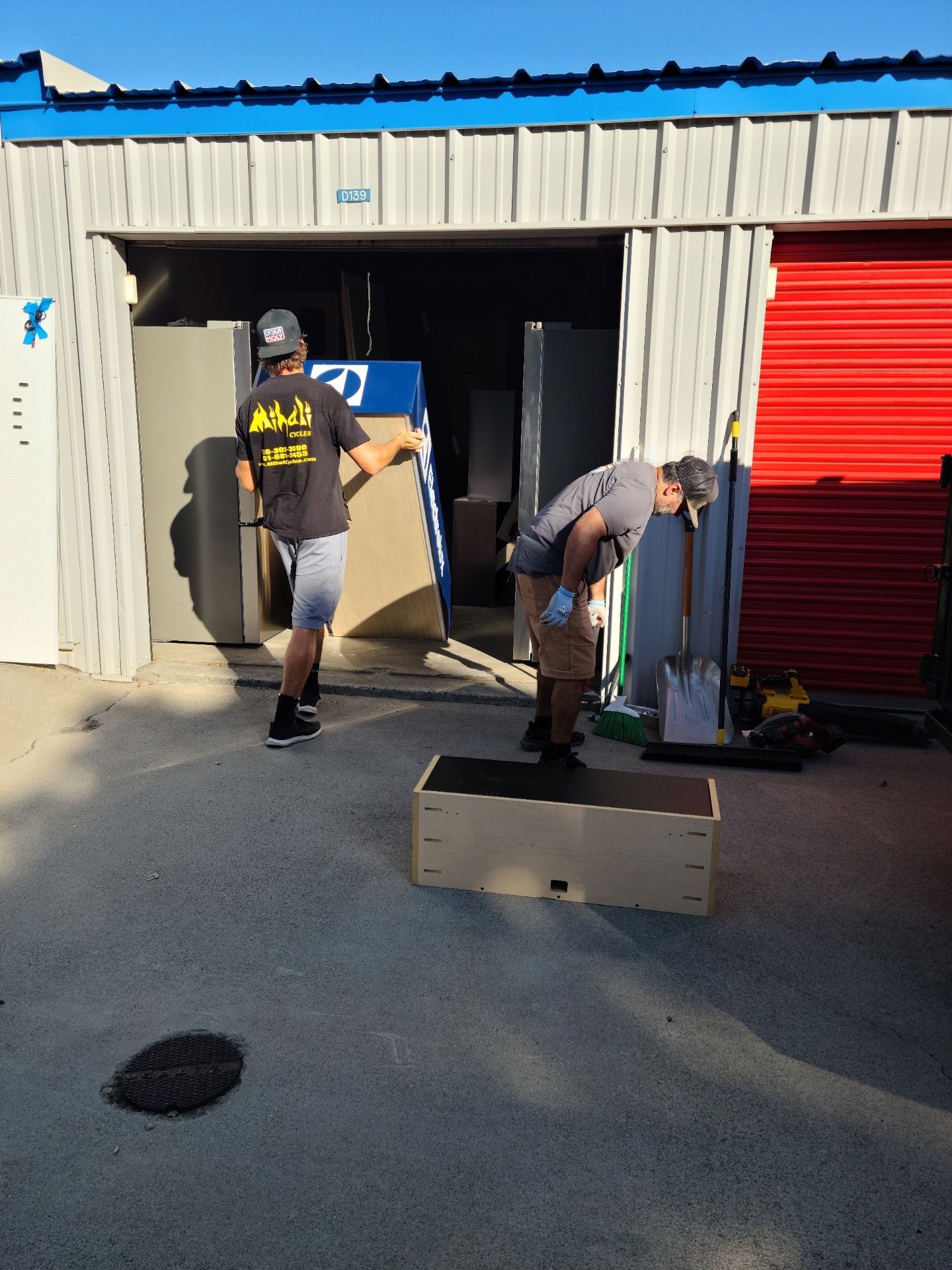 Two people loading a storage unit. One carries a large board, the other handles a box. Sunny day.