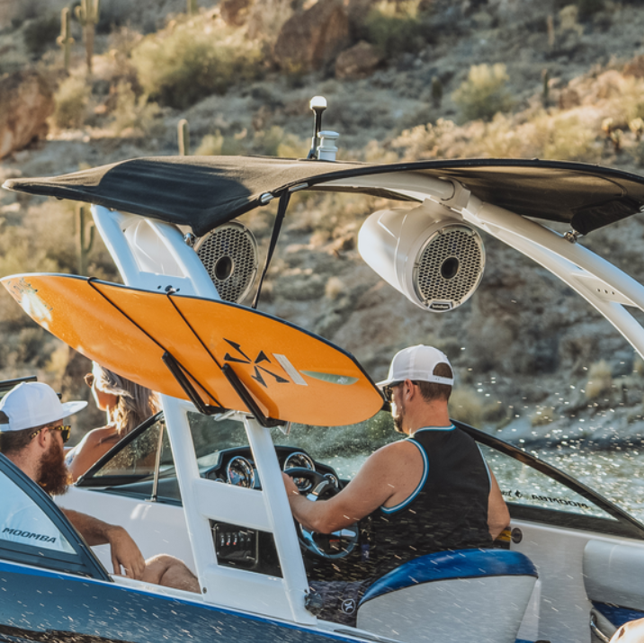 People in a boat on a lake, featuring a wakeboard mounted on the tower under a canopy, with rocky terrain in the back.