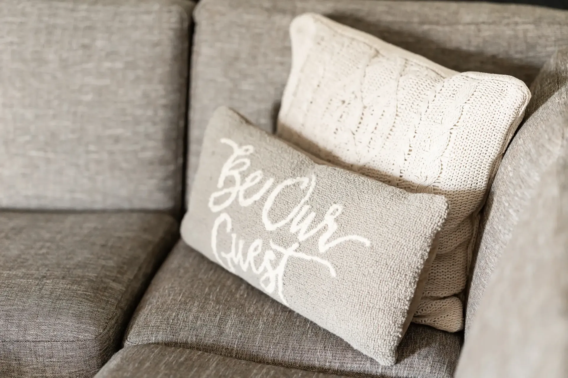 Two pillows are sitting on a couch and one of them says `` be our guest ''.