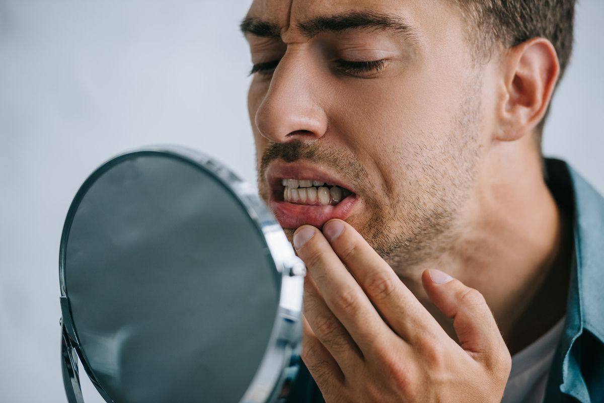 Man in a green collared shirt, standing and holding a mirror to check his teeth and gums