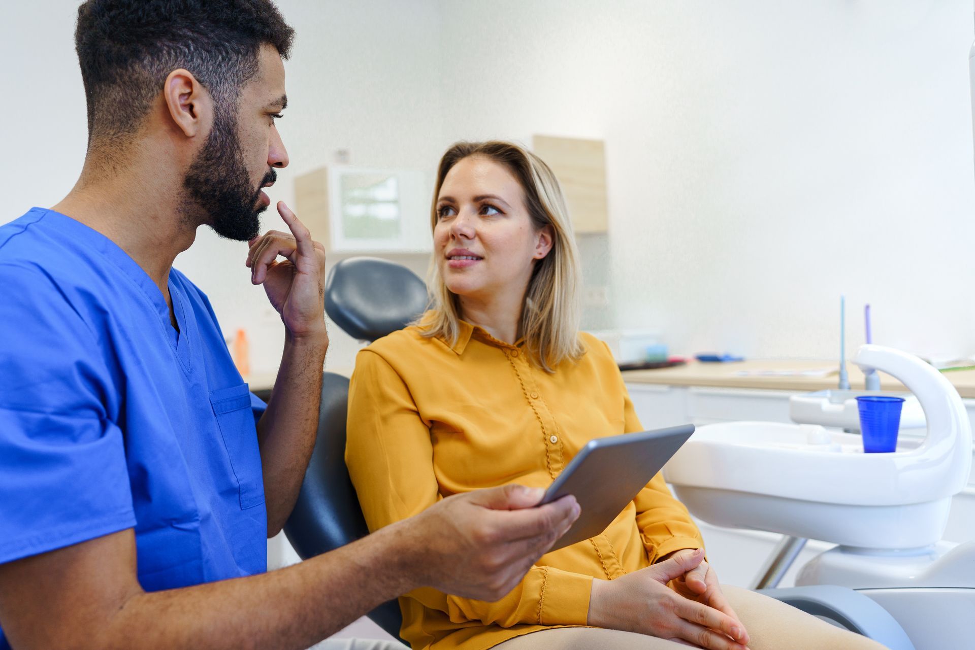 Dental professional discussing treatment with a patient in a clinic, holding a tablet beside dental equipment.