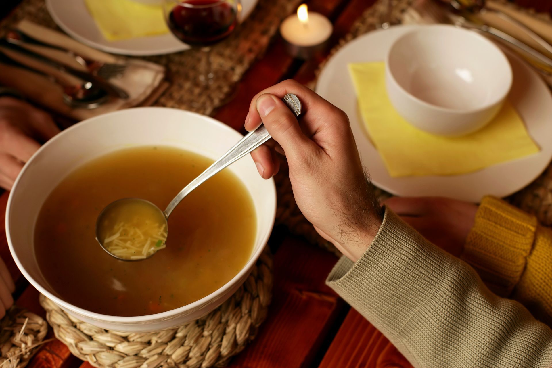 Hand stirring soup in a large white bowl beside an empty cup on a table with a candle and napkins