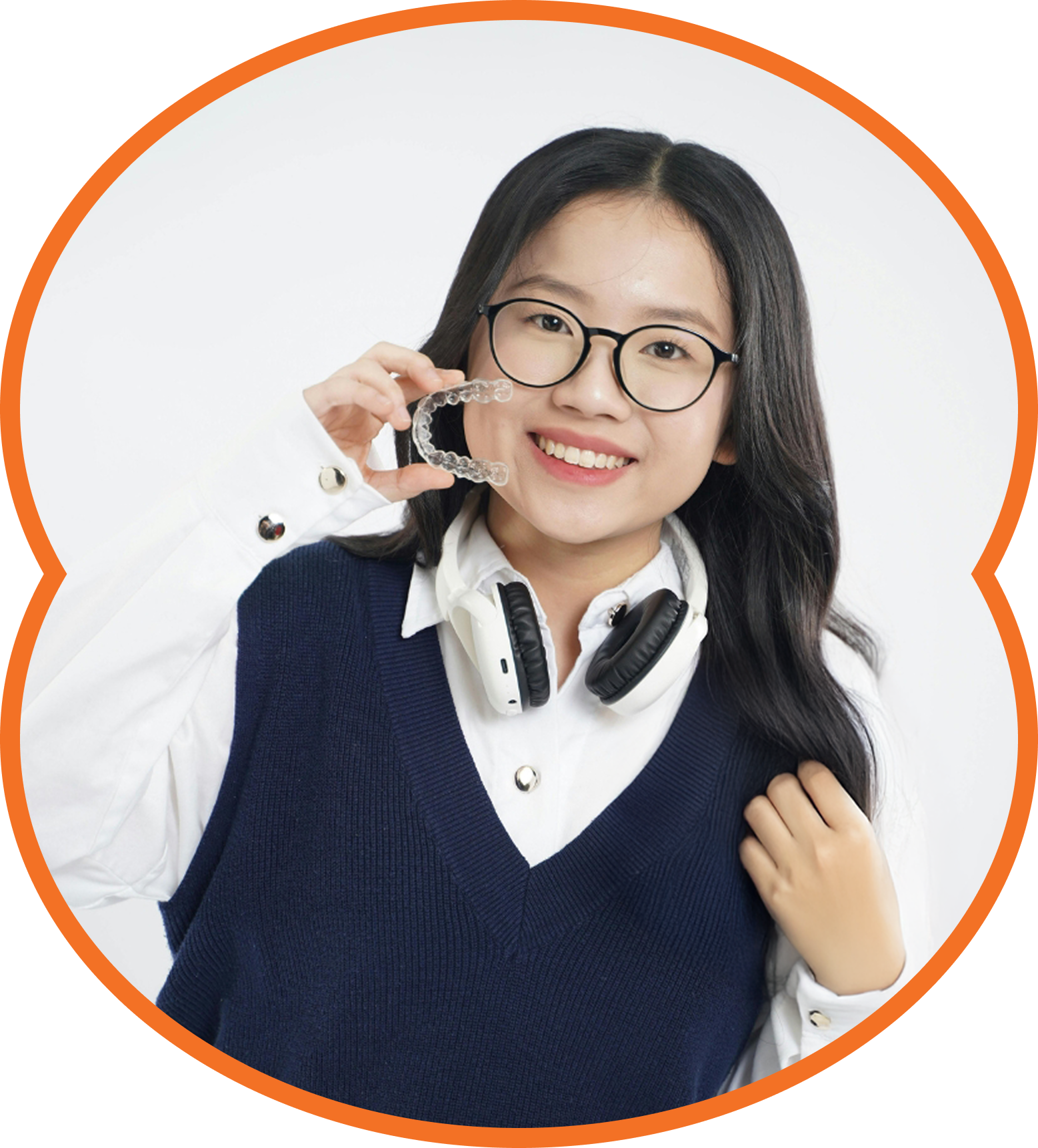 Smiling woman holding eyeglasses, wearing a navy vest and headphones around her neck on a white background.