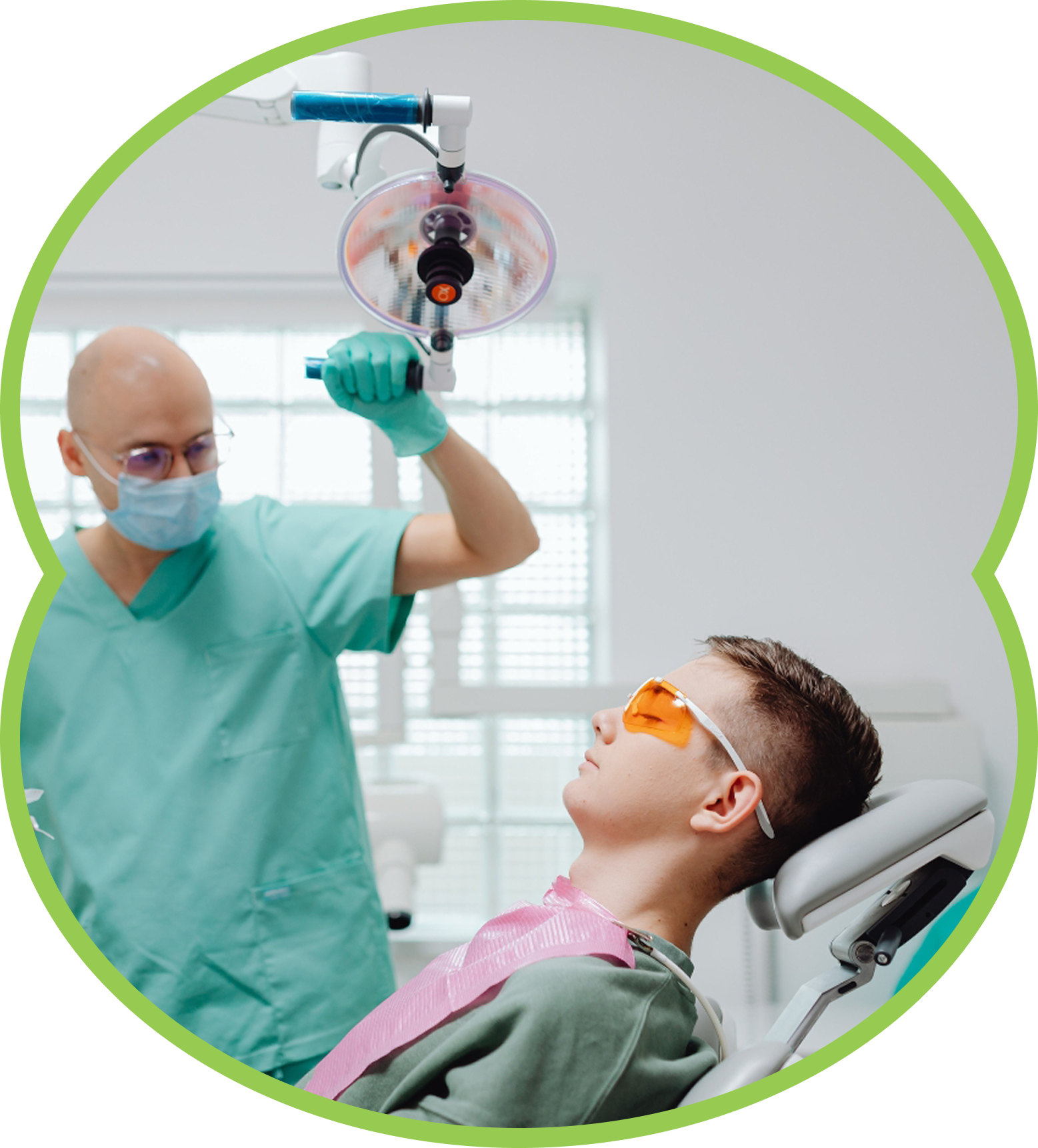 Dentist in green scrubs examines a reclining patient wearing orange glasses in a clinic