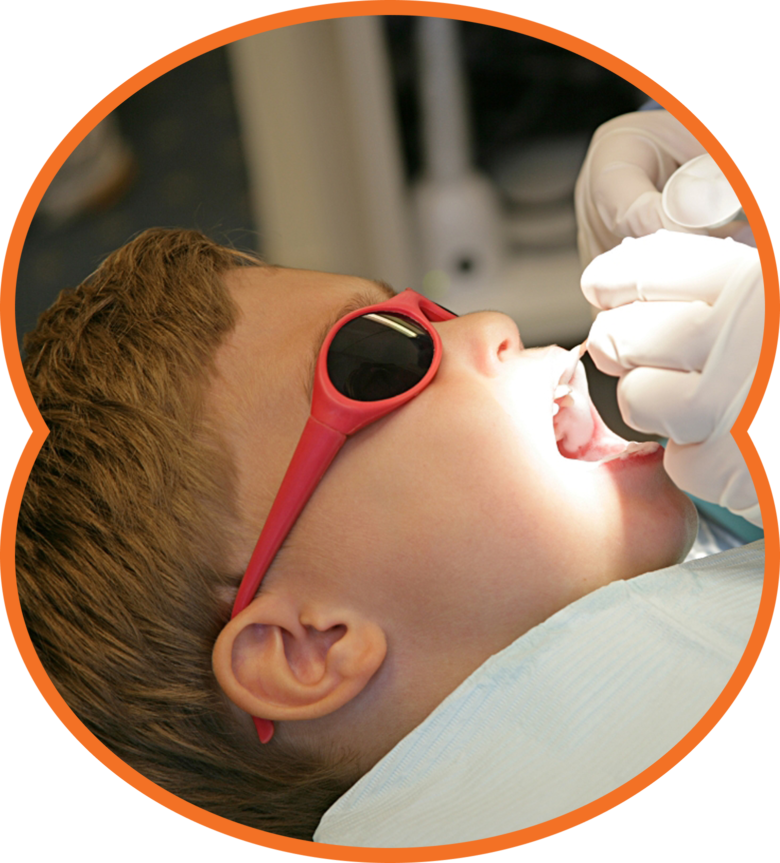 Child wearing red sunglasses during a dental exam with mouth open and dentist's gloved hand visible