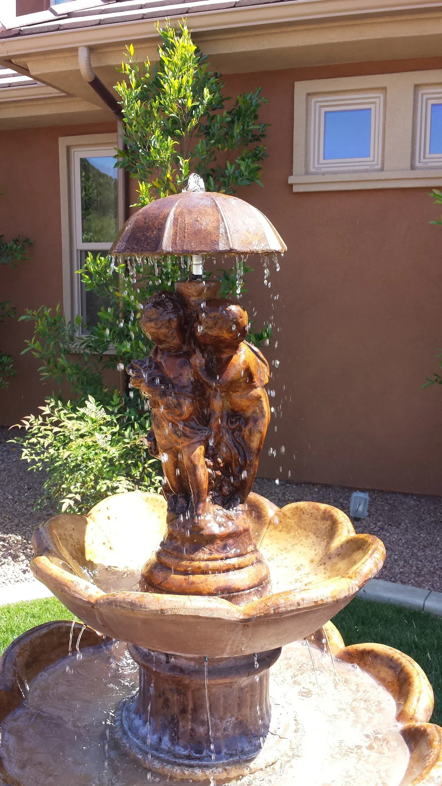 Water fountain with brown figures under an umbrella, water cascading into a flower-shaped basin, set outdoors.