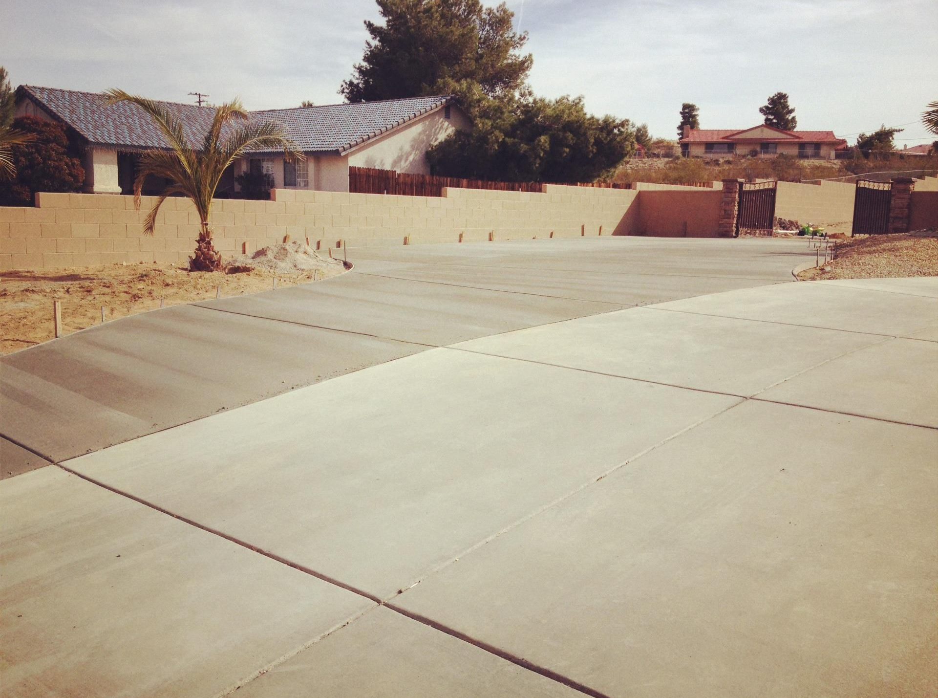 Concrete driveway leading to a house with a wall and palm tree. Blue roof visible.