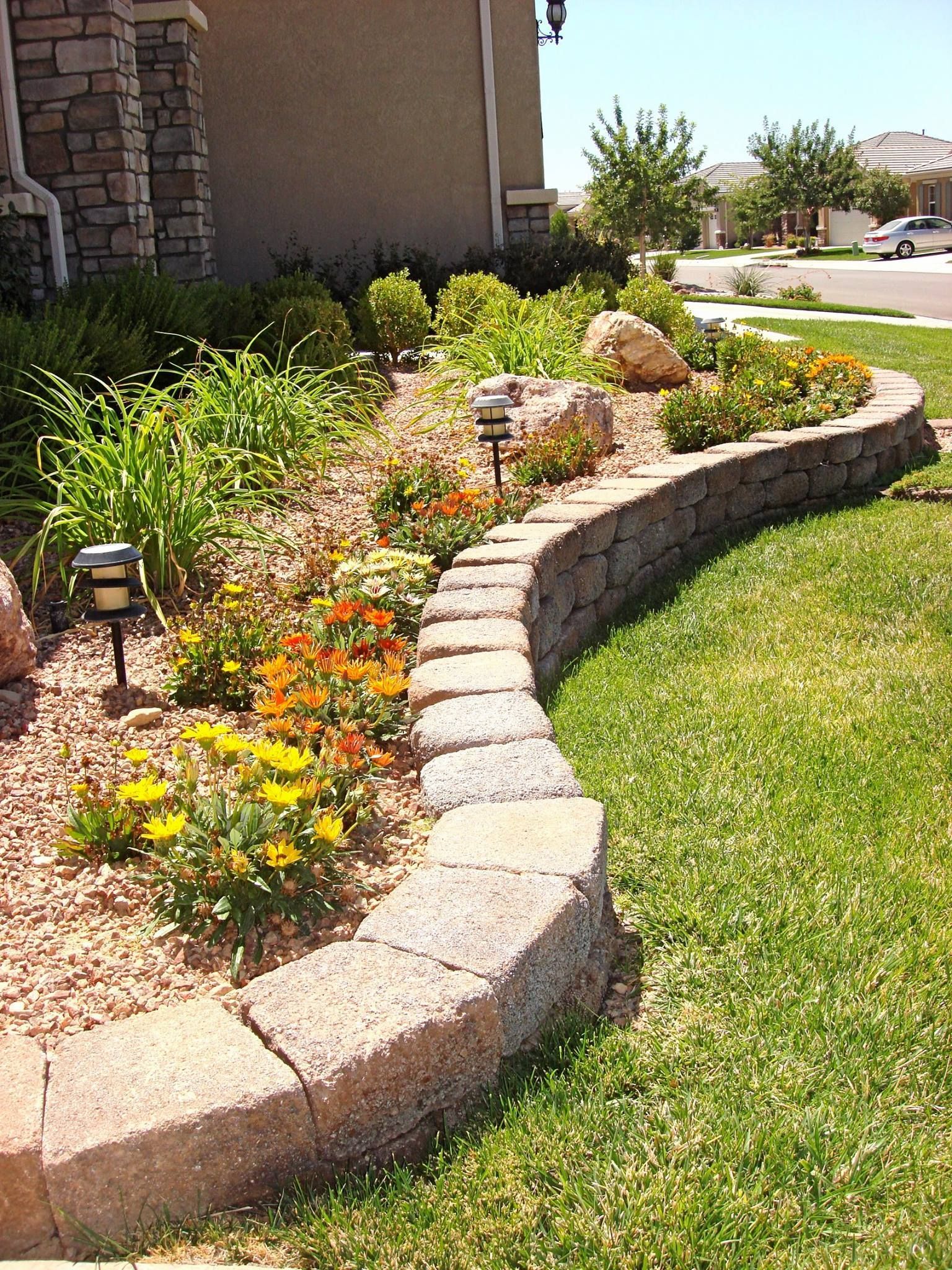 Stone-edged garden bed with colorful flowers, small solar lights, and green grass in front of a house.