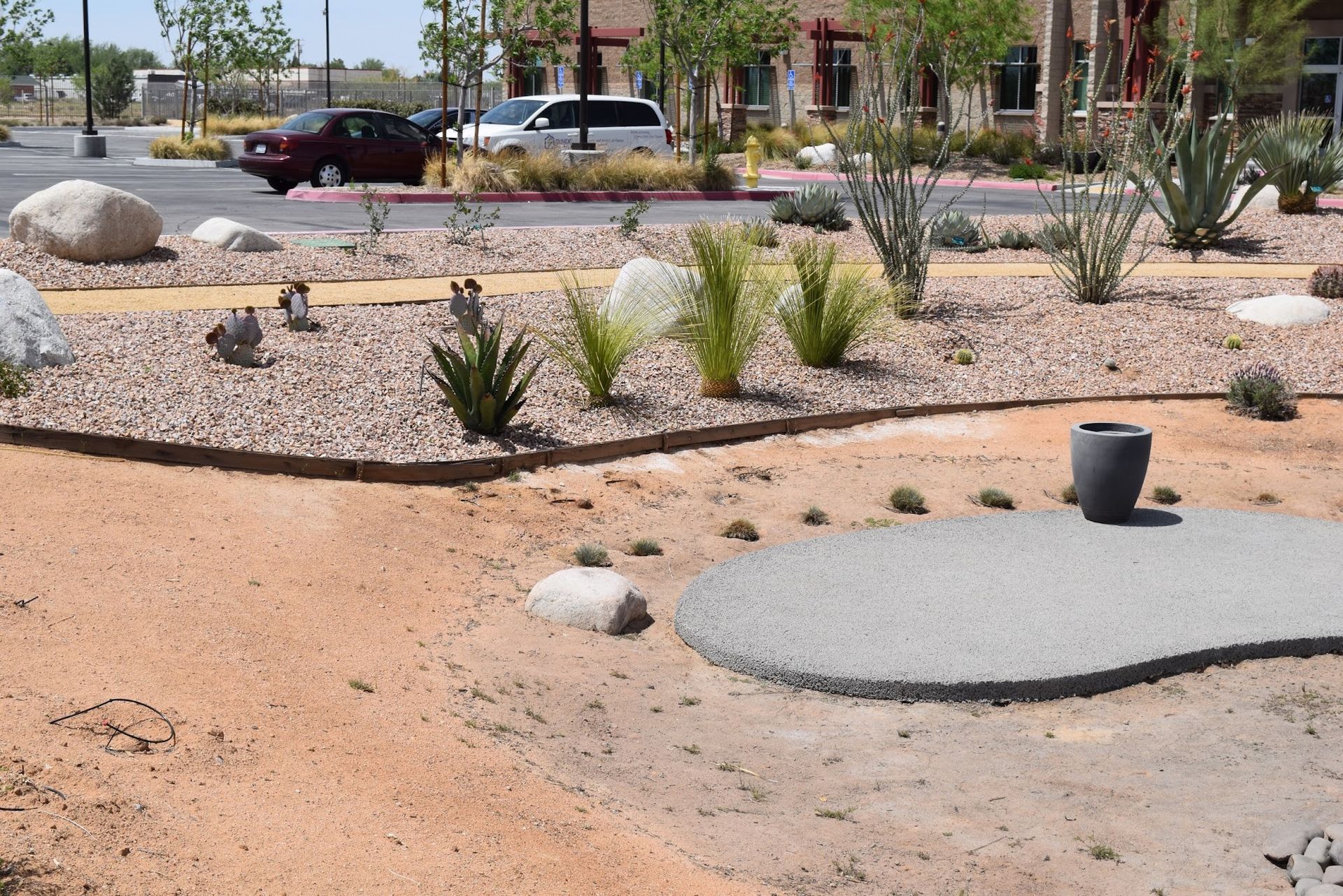 Desert landscaping with gravel, plants, rocks, and a circular paved area. Cars and a building are in the background.