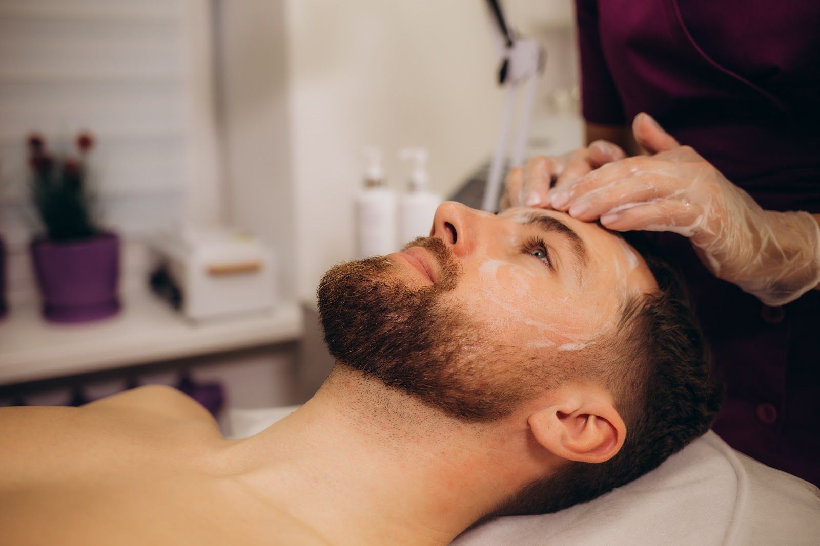 Man receiving a facial massage in a spa treatment room, with a hand applying cream to his forehead.