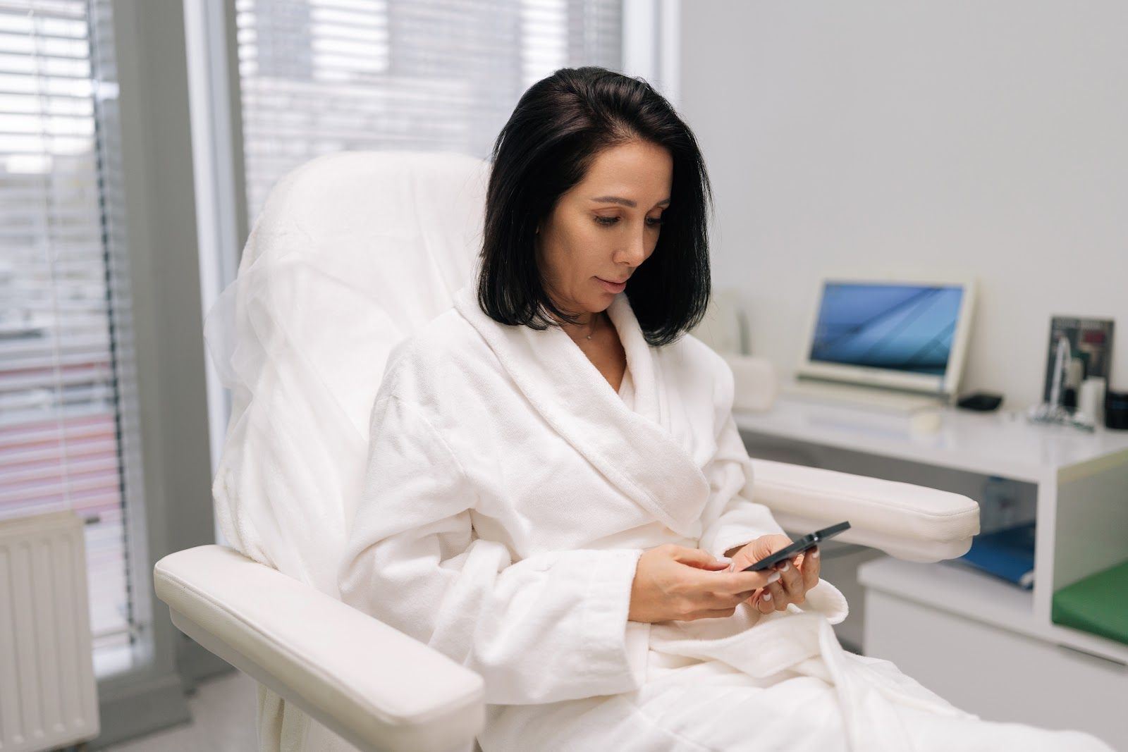 Woman in a white robe seated in a clinic chair, looking at her phone in a bright room