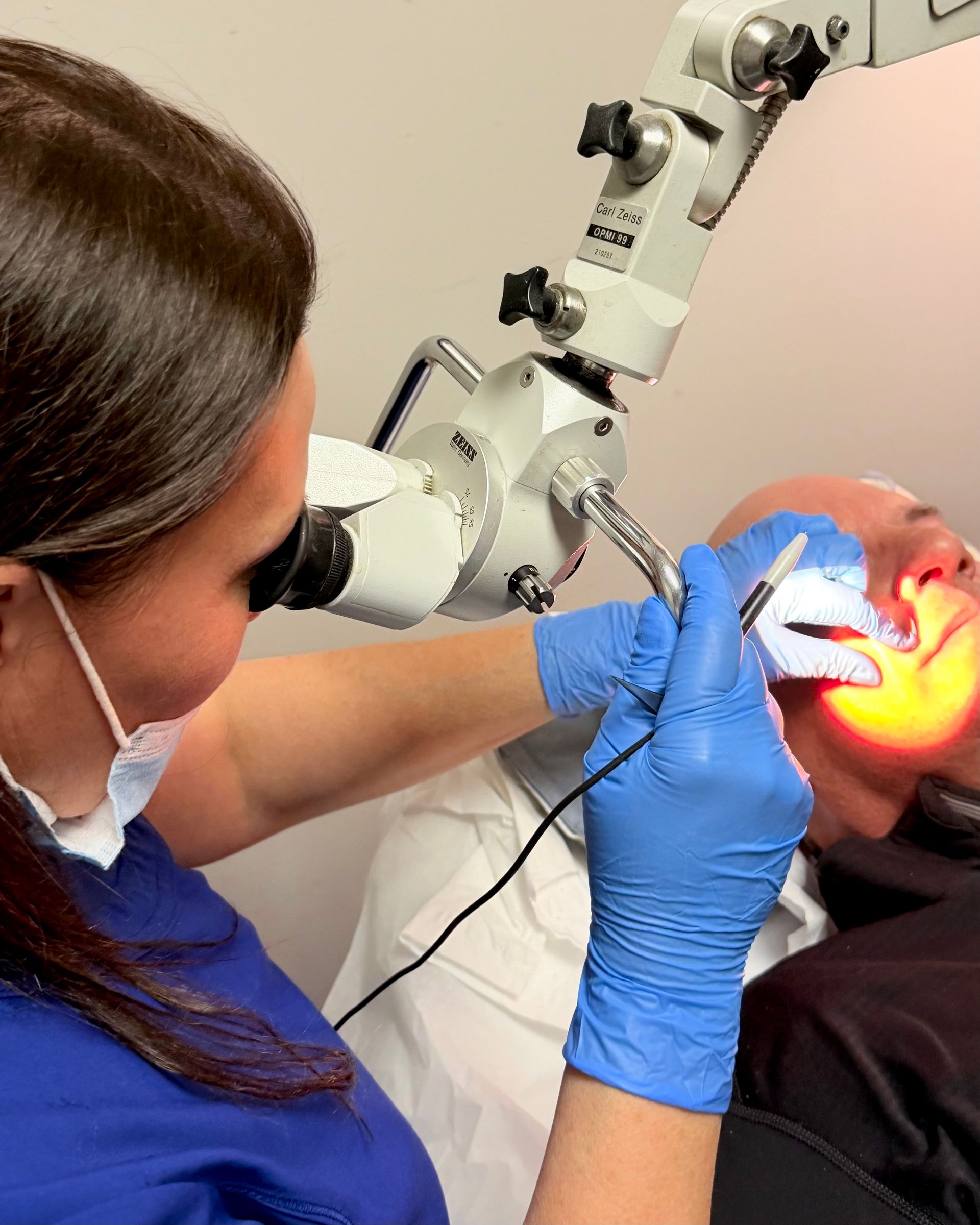 Dentist using a microscope and tools to examine a patient’s mouth under bright light