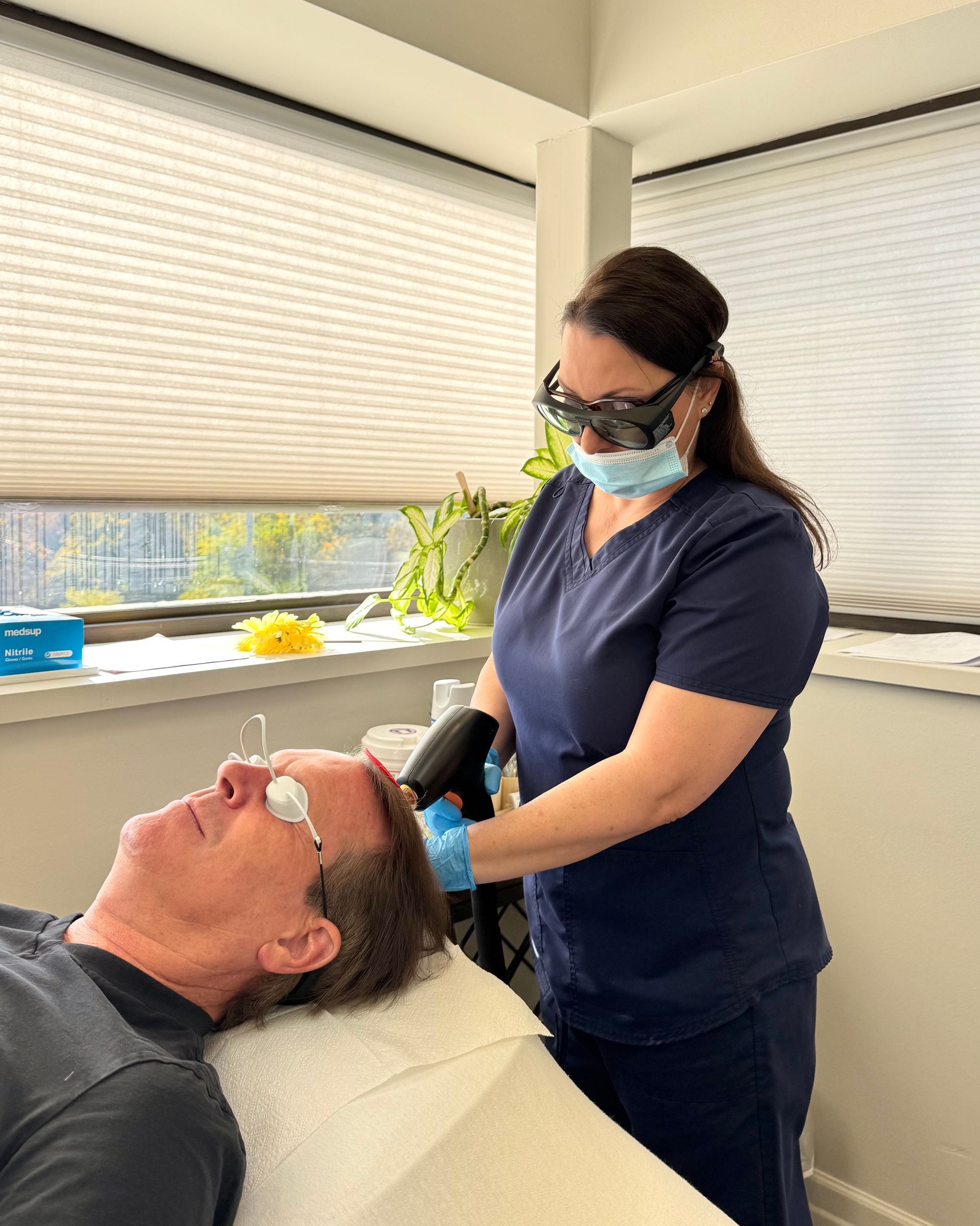 Dentist wearing mask examines a patient reclined in a chair by a bright window.
