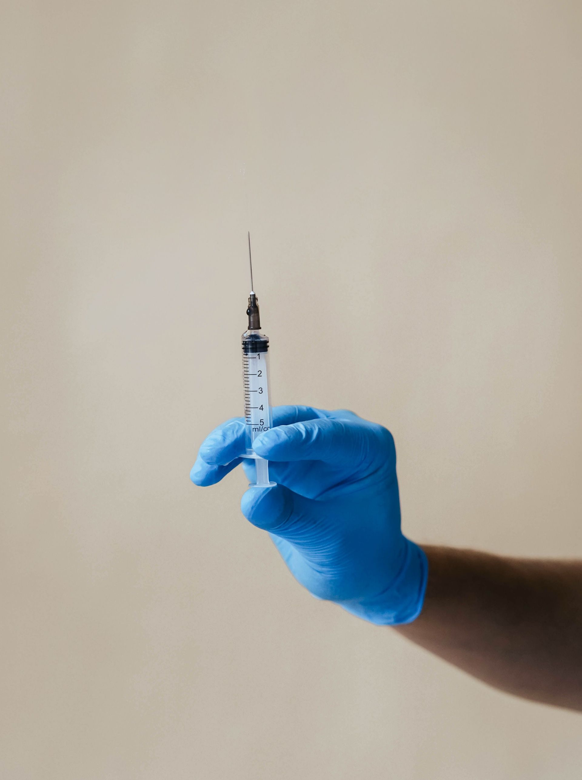 Gloved hand holding a syringe upright against a plain beige background