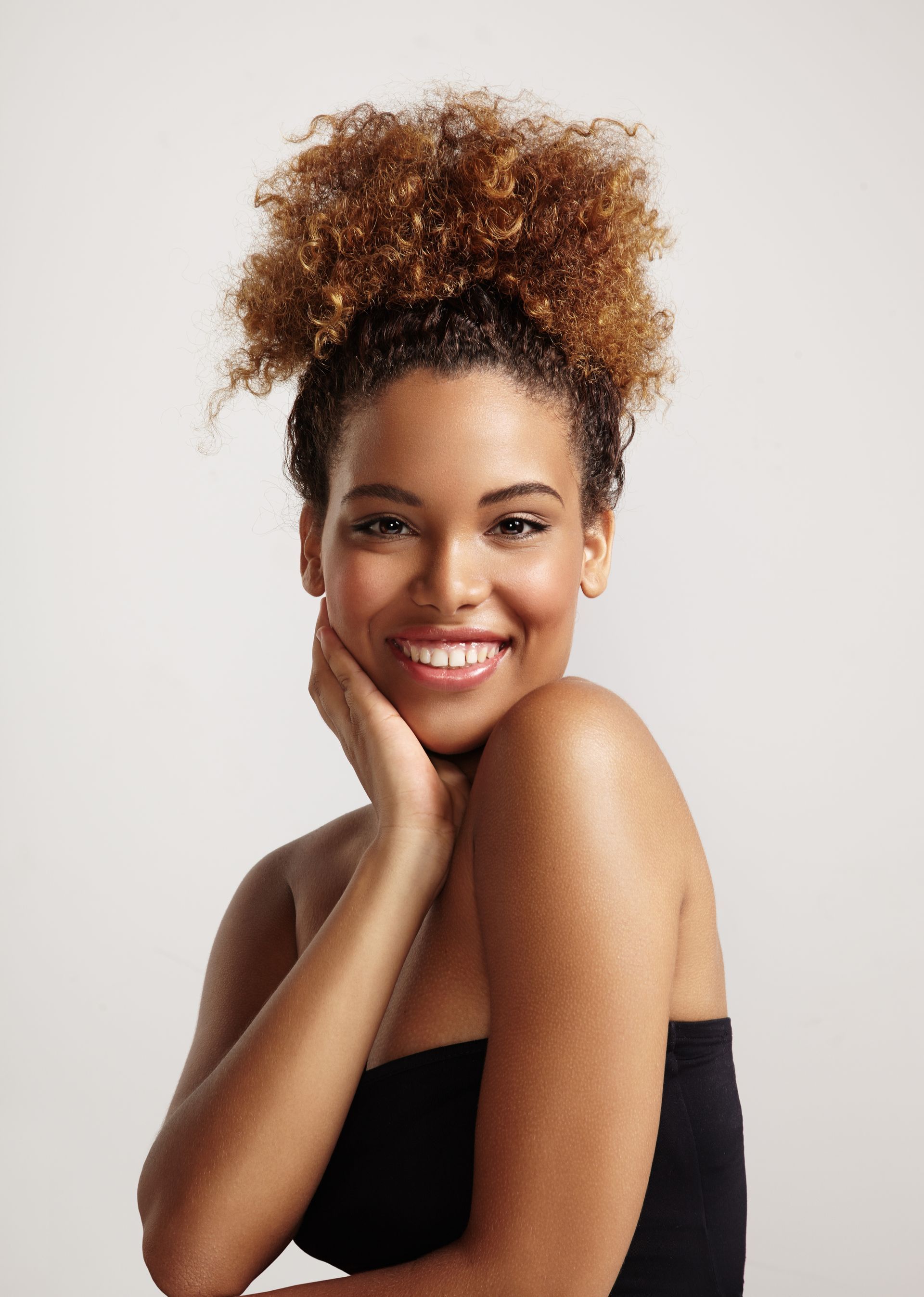 Smiling person in a black top with curly hair, resting chin on hand against a light background