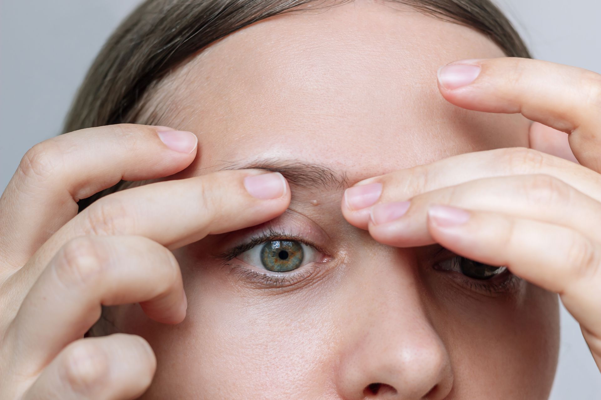 Close-up of hands shaping eyebrows on a person’s face, with one green eye visible.