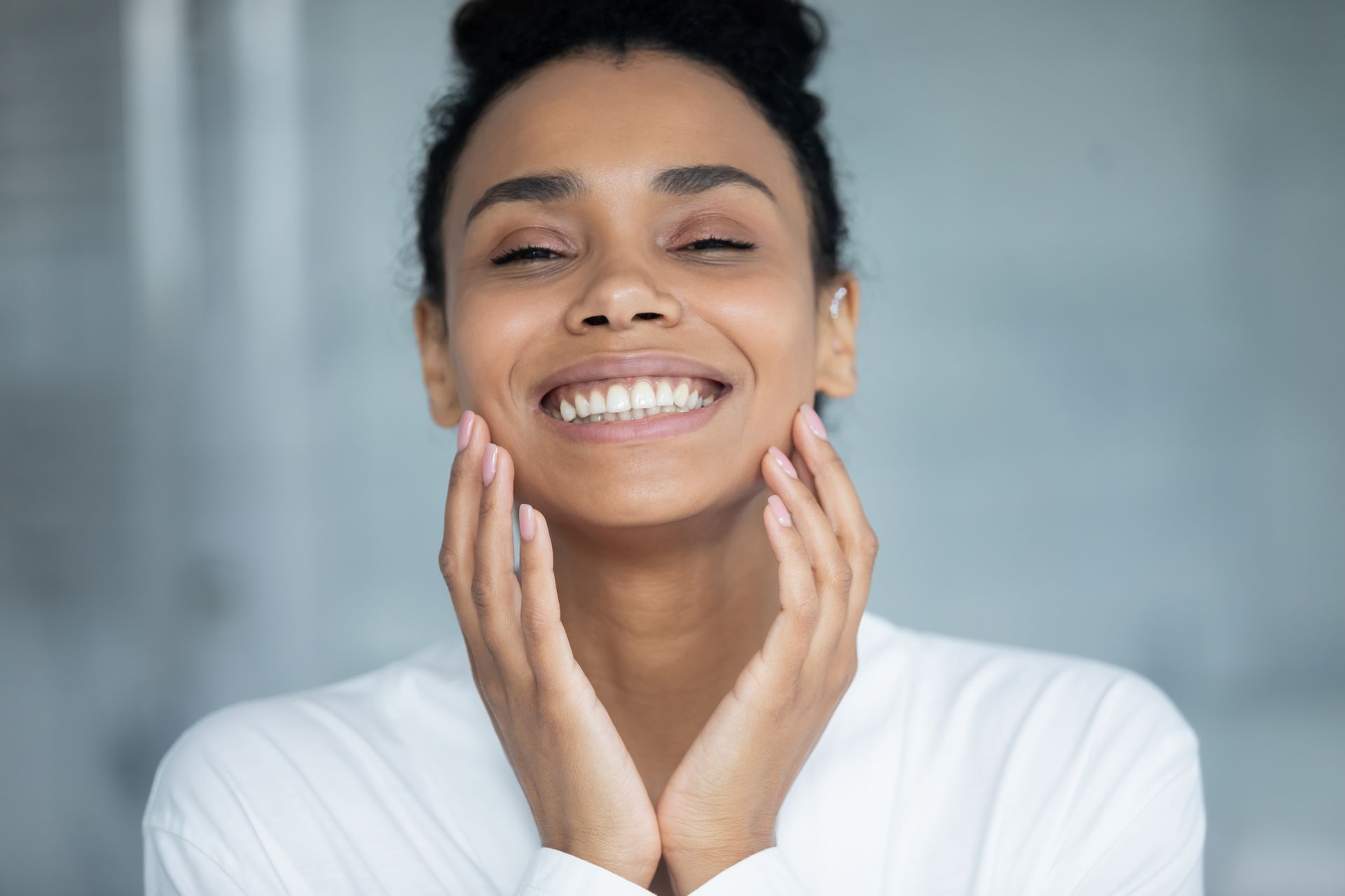 Smiling person in a white top gently touching their cheeks against a soft gray background