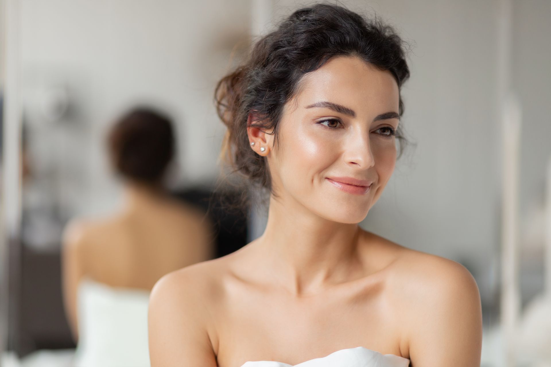 Woman with dark hair in a white top, smiling in a softly lit room with a blurred mirror in the background